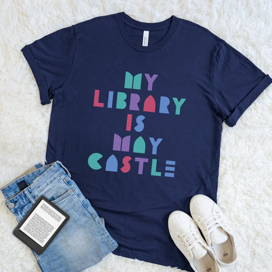 Navy book lover T-shirt with bright My Library Is My Castle typography styled with jeans and an e-reader.