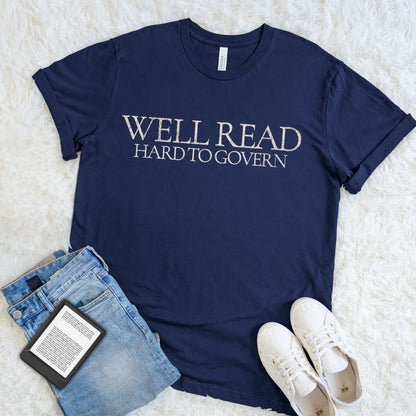 A navy blue graphic t-shirt with the text 'Well read hard to govern' in white serif letters, styled in a flat lay on a white faux fur background with blue jeans, white sneakers, and a book.