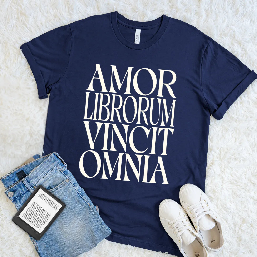 A navy blue bookish graphic t-shirt with the phrase 'AMOR LIBRORUM VINCIT OMNIA' printed in large white serif letters, styled in a flat lay on a white faux fur background with blue jeans, a book, and white sneakers.