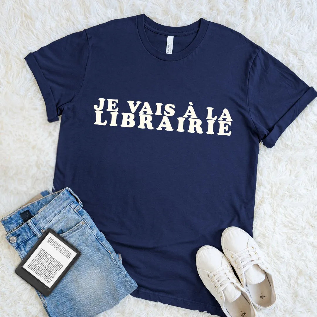 Navy Je Vais à La Librairie French bookstore T-shirt with bold cream retro lettering, styled with jeans, white sneakers, and an e-reader