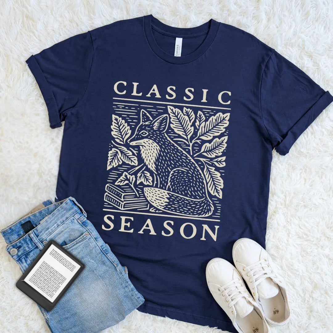 Navy Classic Season T-shirt with an ivory woodcut fox, stacked books, and leafy woodland branches, styled with jeans and an e-reader
