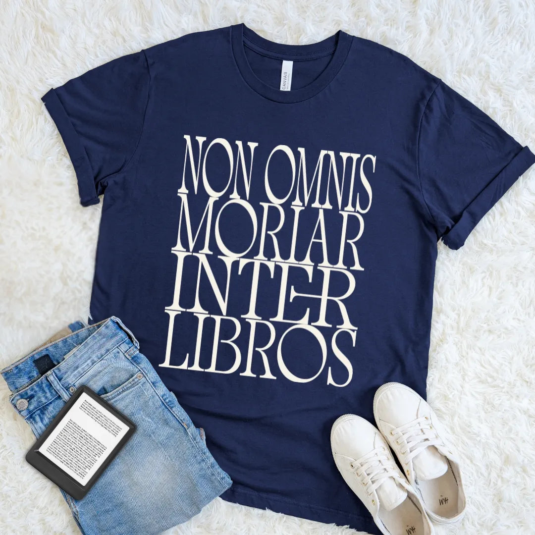 A navy blue graphic t-shirt with the with bold vintage typography 'NON OMNIS MORIAR INTER LIBROS' printed in large white serif letters, styled in a flat lay on a white faux fur background with blue jeans, a book, and white sneakers.