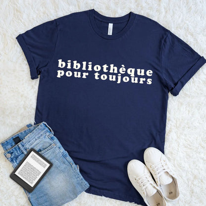 Navy Francophile reader tee with cream Bibliothèque Pour Toujours lettering beside folded jeans, an e-reader, and white shoes