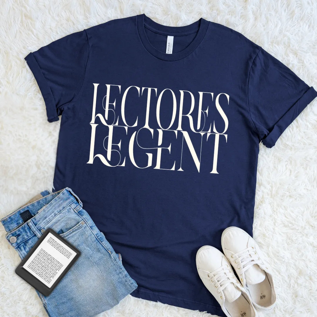 A navy blue graphic reader t-shirt with the words 'LECTORES LEGENT' printed in white stylized lettering, styled in a flat lay with blue jeans, a book, and white sneakers on a white faux fur background.