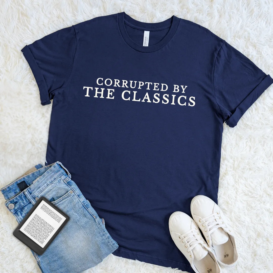 A navy blue graphic t-shirt with the text 'CORRUPTED BY THE CLASSICS' in white serif letters, styled in a flat lay with blue jeans, a book, and white sneakers on a white faux fur background.