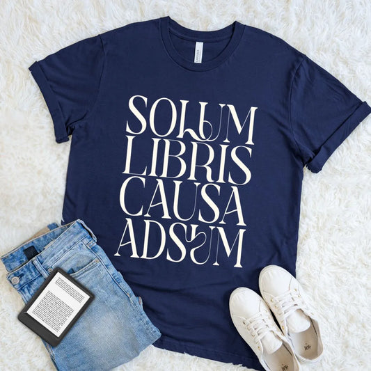 A navy blue book lover graphic t-shirt with the text 'SOLUM LIBRIS CAUSA ADSUM' in white stylized lettering, styled in a flat lay with light-wash denim jeans, white sneakers, and a book on a white faux fur background.