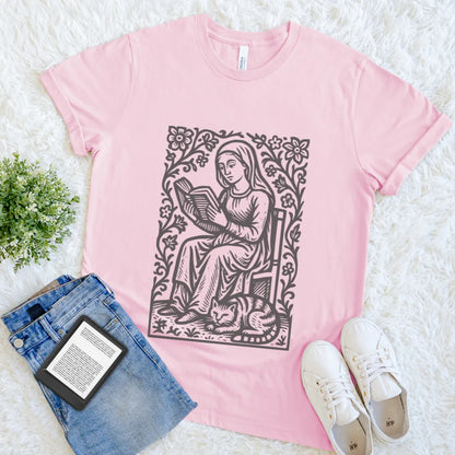 Pink t-shirt with a graphic design of a woman reading a book, surrounded by flowers, on a white surface with jeans and sneakers.