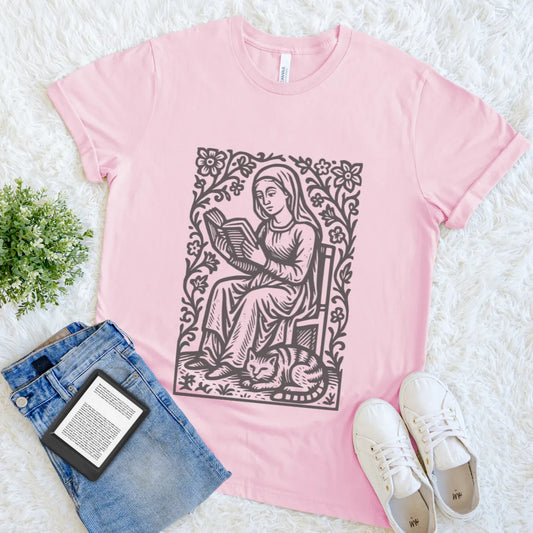 Pink t-shirt with a graphic design of a woman reading a book, surrounded by flowers, on a white surface with jeans and sneakers.