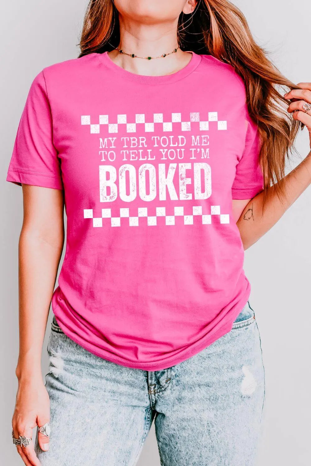woman  wearing pink soft cotton shirt with bold white text reading “My TBR told me to tell you I’m BOOKED” in distressed font