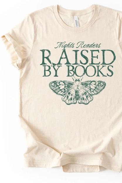 Flat lay of cream-colored bookish t-shirt with green vintage-style text reading “Raised by Books” and a detailed butterfly illustration, creating a soft book lover aesthetic.
