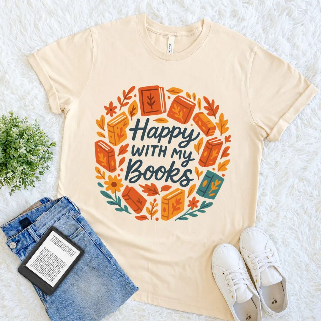 A soft cream bookish graphic T-shirt laid flat on a white textured surface, surrounded by styled props including blue jeans, white sneakers, a small plant, and an open book. The shirt features a circular autumn-themed book illustration with the phrase “Happy with my books” in dark lettering.