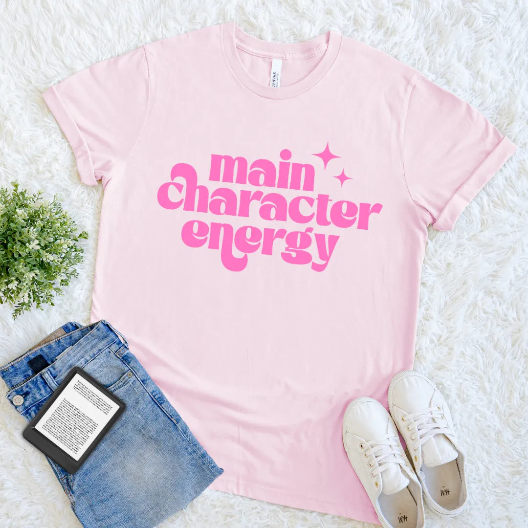 Pink t-shirt with 'main character energy' text, paired with jeans and sneakers on a white surface.