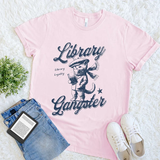 Soft Pink cotton t-shirt with cat and book illustration and a 'Library Gangster' quote on a white surface with denim shorts and sneakers.