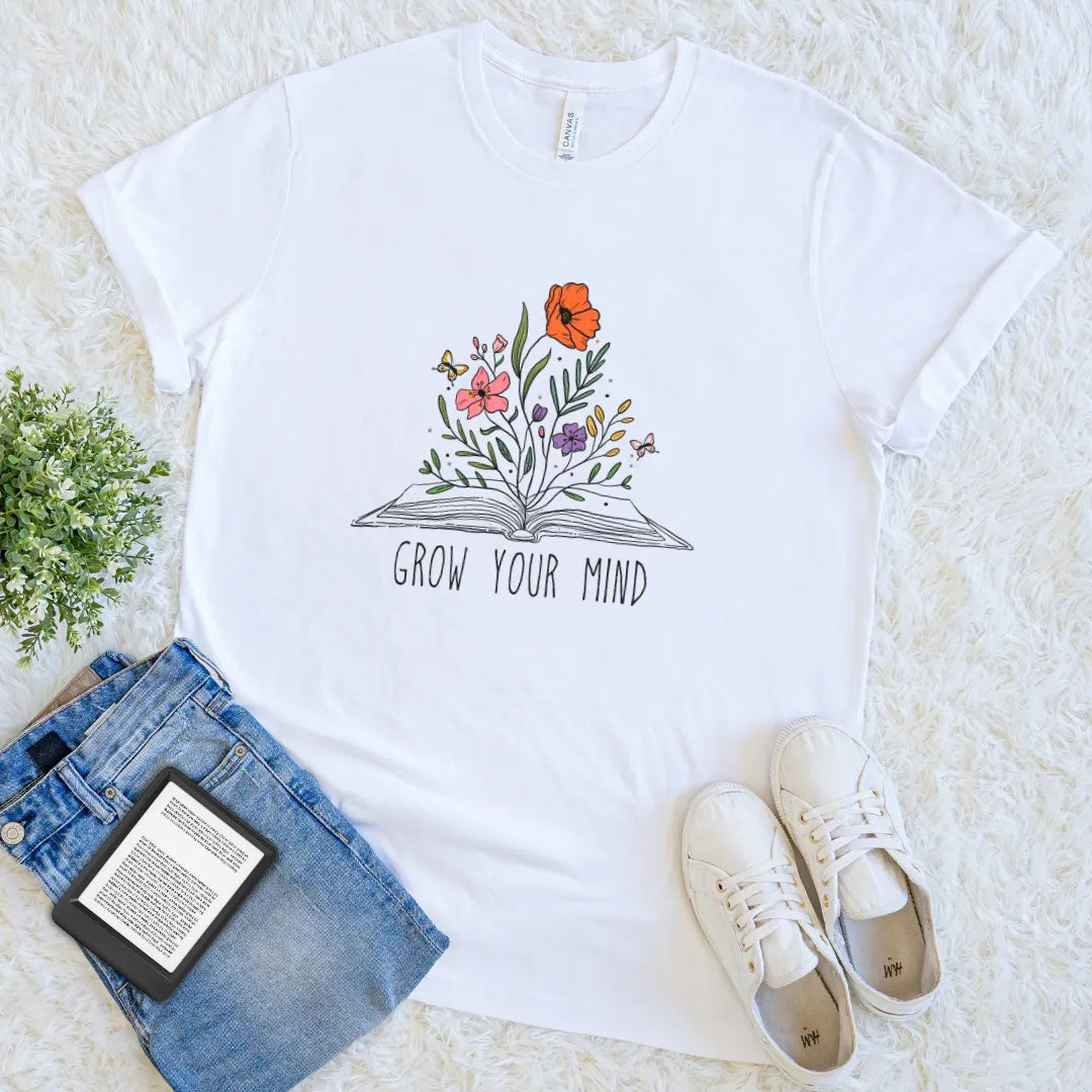 White t-shirt with floral book design and 'Grow Your Mind' text, placed on a light surface with jeans and sneakers.