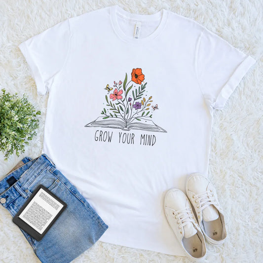 White t-shirt with floral book design and 'Grow Your Mind' text, placed on a light surface with jeans and sneakers.
