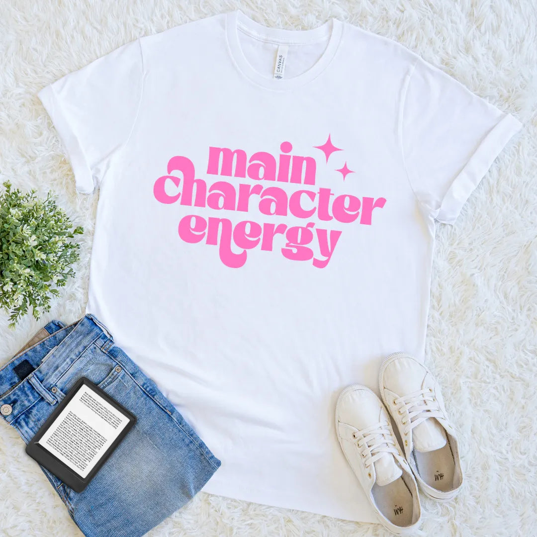 White t-shirt with pink text 'main character energy', paired with denim shorts and white sneakers on a light background.
