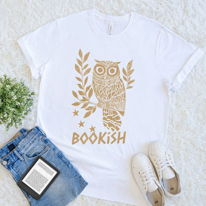White t-shirt with gold owl design and 'bookish' text, placed on a light surface with jeans and sneakers.