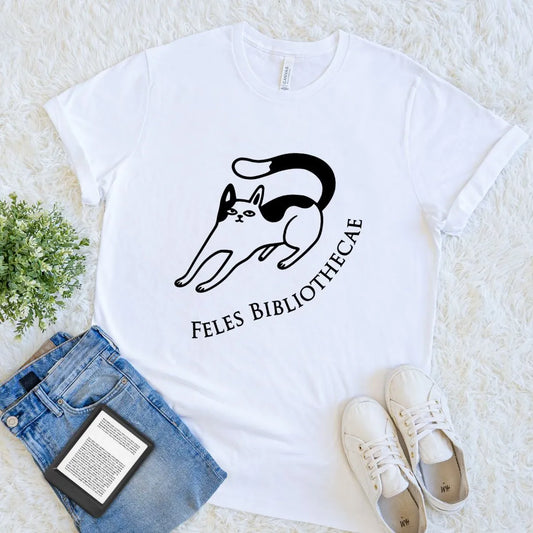 A white short-sleeve graphic T-shirt for readers laid flat on a white textured rug, styled with folded blue jeans, a small green plant, and white sneakers. The shirt has a black line drawing of a cat and the text 'FELES BIBLIOTHECAE' printed on the front.