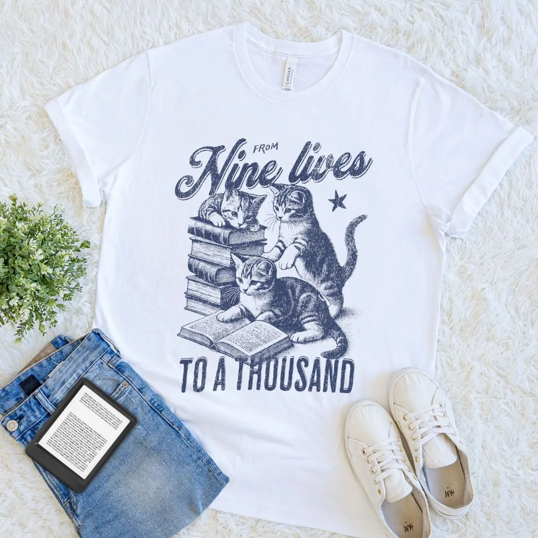 Flat lay of white soft cotton cat-themed book lover tee with stacked books and cozy motif