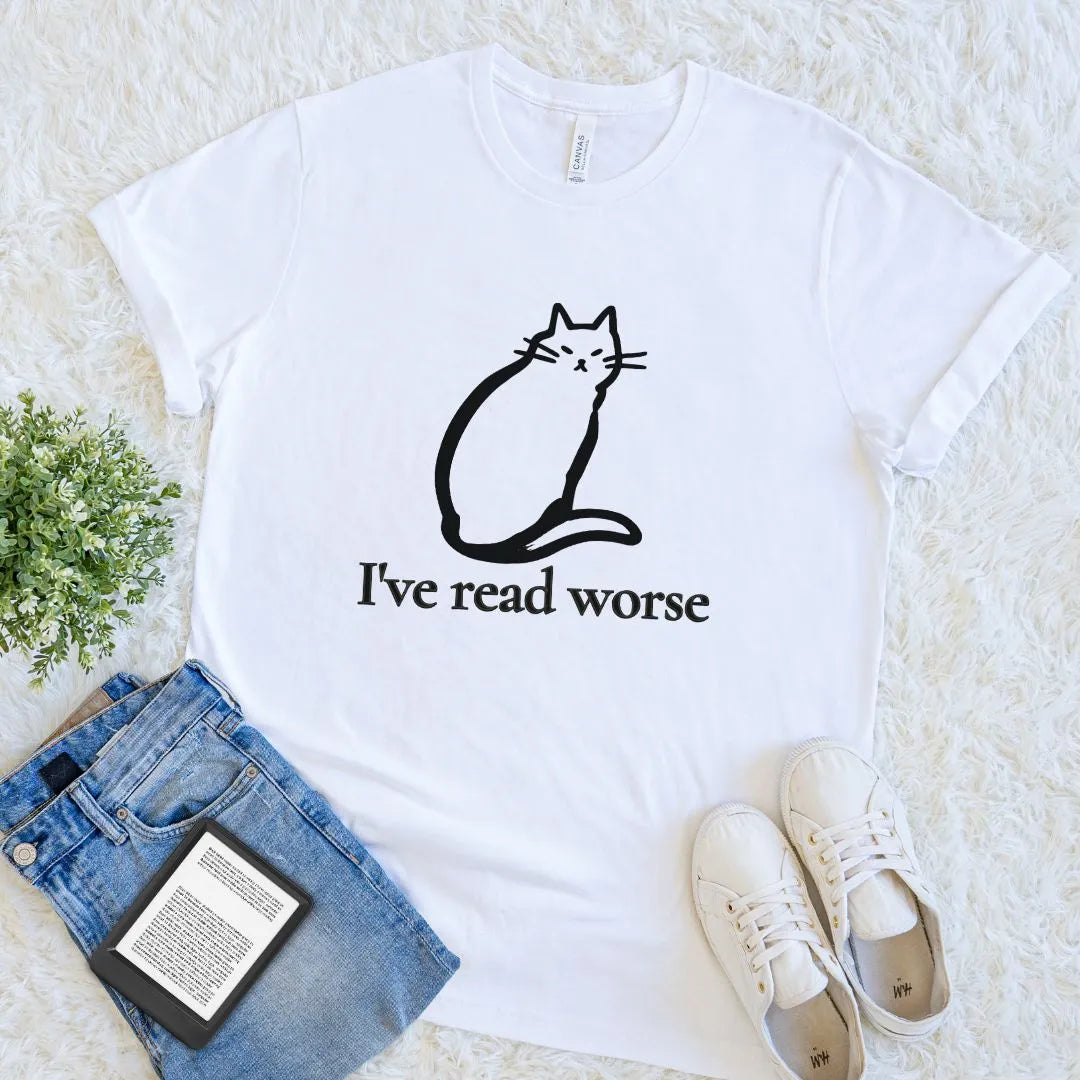 A white short-sleeve graphic T-shirt laid flat on a white textured rug, styled with folded blue jeans, a small green plant, a book, and white sneakers. The shirt has a black line drawing of a cat and the witty reading slogan 'I've read worse' printed on the front.