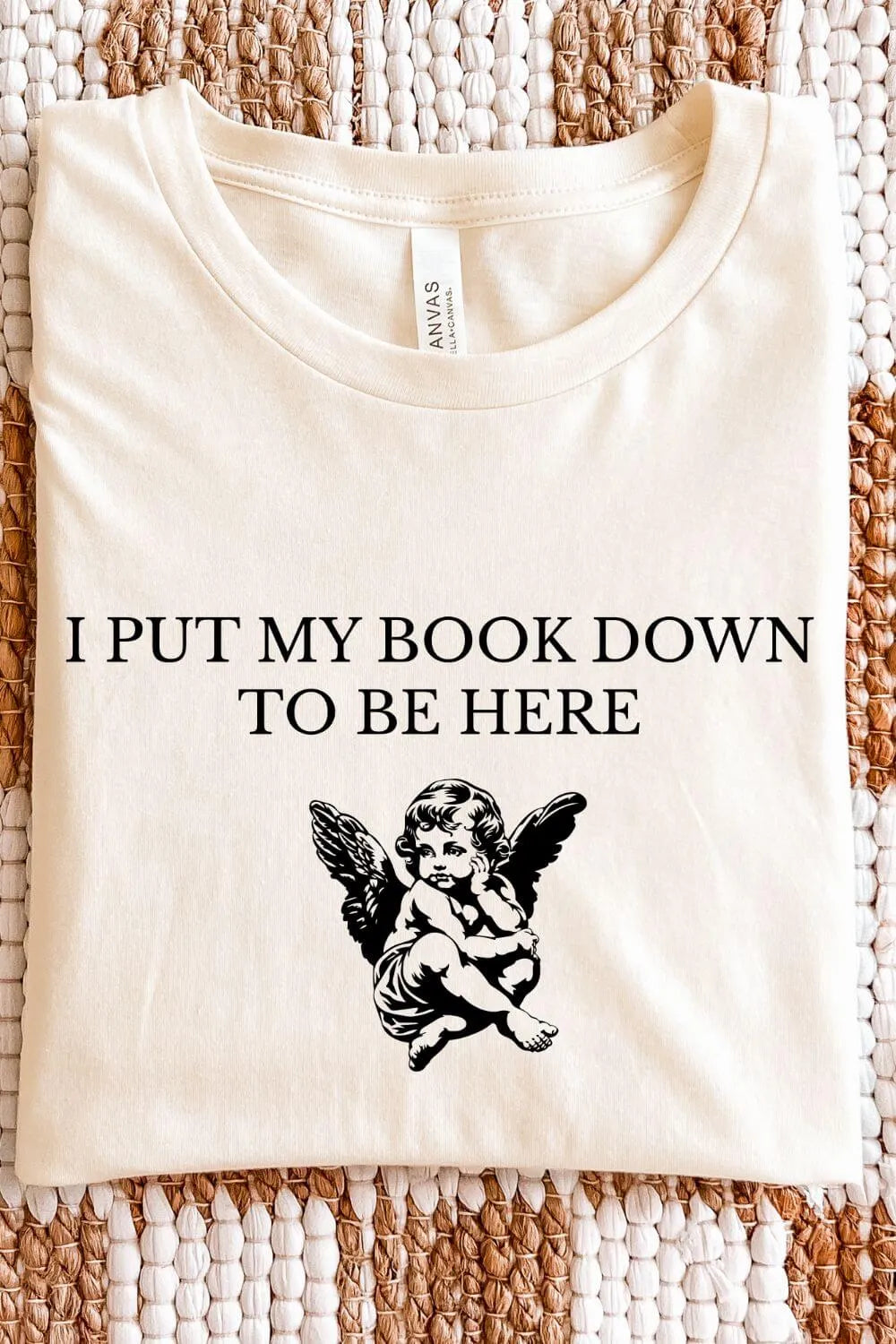 A soft cotton cream-colored t-shirt featuring a vintage cherub illustration and the phrase “I Put My Book Down to Be Here,” styled in an angelcore bookish aesthetic.