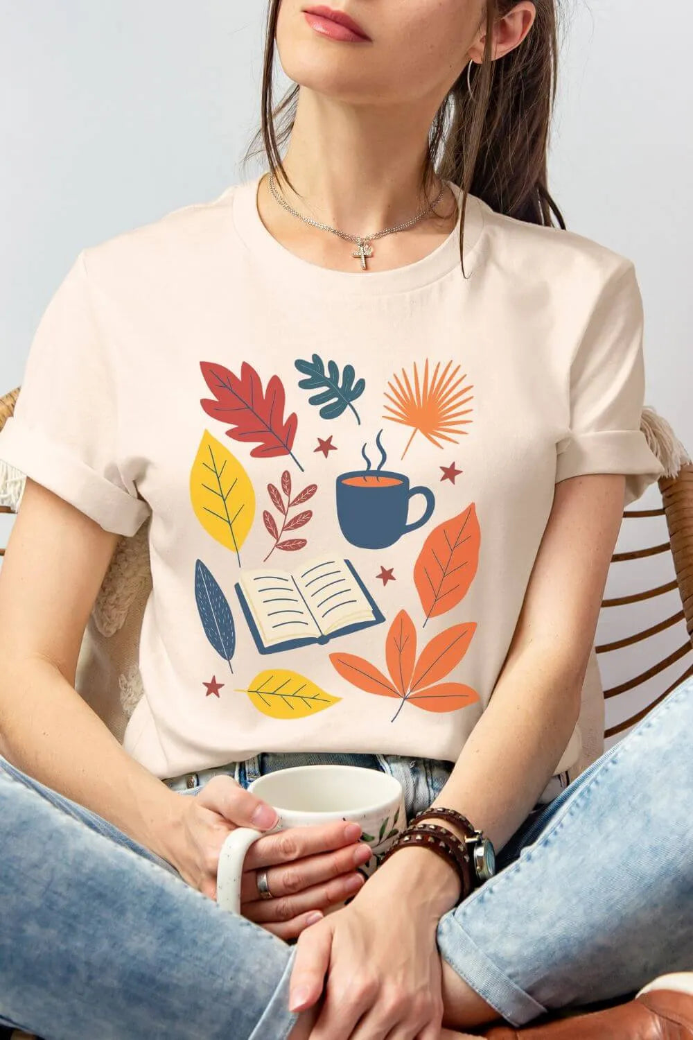 Person wearing a cotton soft cream t-shirt  featuring an illustrated open book, coffee cup, and colorful fall leaves in warm autumn tones, styled as a cozy reading outfit with denim