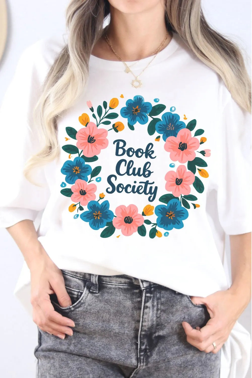A white t-shirt worn by a model featuring a colorful floral wreath design with the words ‘Book Club Society’ in the center.”