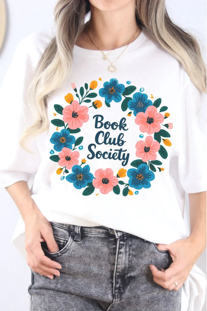 A white t-shirt worn by a model featuring a colorful floral wreath design with the words ‘Book Club Society’ in the center.”