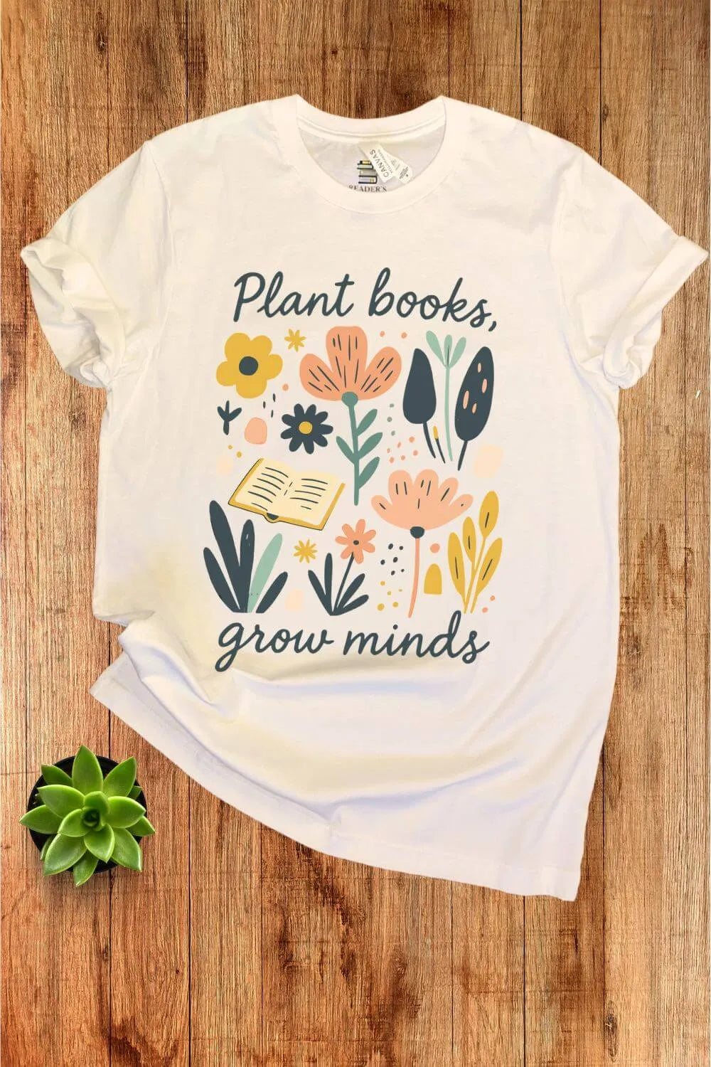 A soft cotton cream  t-shirt with colorful floral and leaf illustrations surrounding an open book and the phrase ‘Plant Books, Grow Minds,’ styled flat on a wooden background