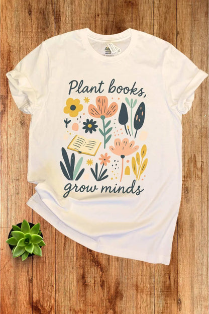 A soft cotton cream  t-shirt with colorful floral and leaf illustrations surrounding an open book and the phrase ‘Plant Books, Grow Minds,’ styled flat on a wooden background