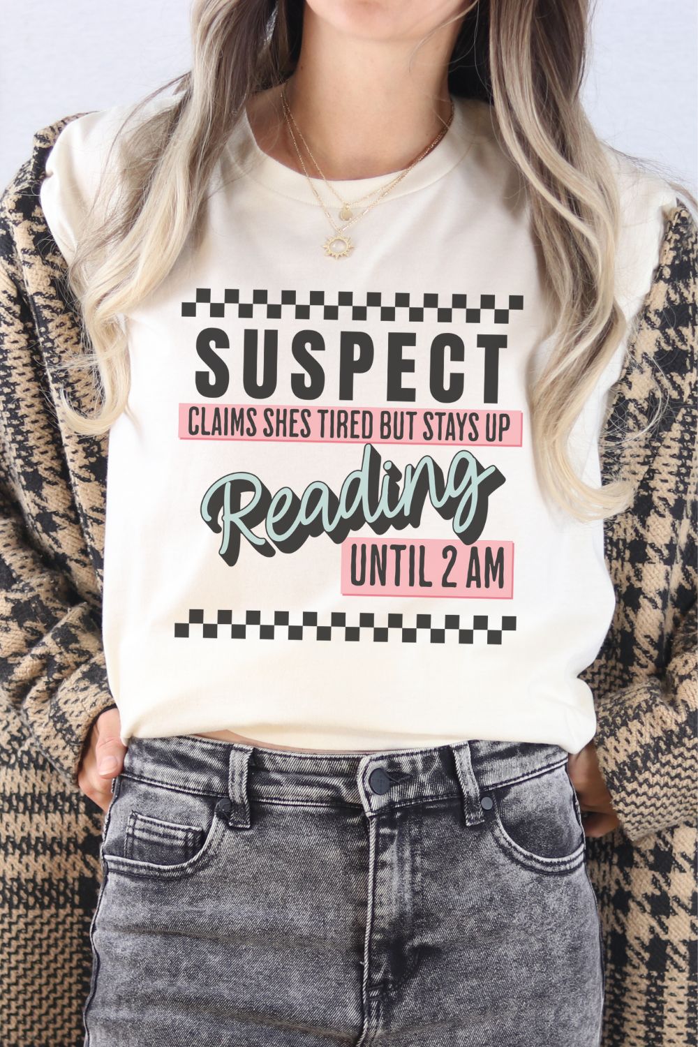 A cream-colored t-shirt worn by a model featuring bold text that reads ‘Suspect: Claims She’s Tired But Stays Up Reading Until 2AM,’ styled casually with jeans.