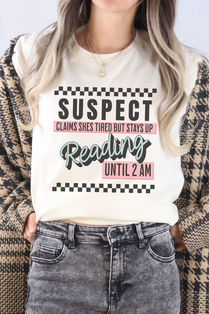 A cream-colored t-shirt worn by a model featuring bold text that reads ‘Suspect: Claims She’s Tired But Stays Up Reading Until 2AM,’ styled casually with jeans.