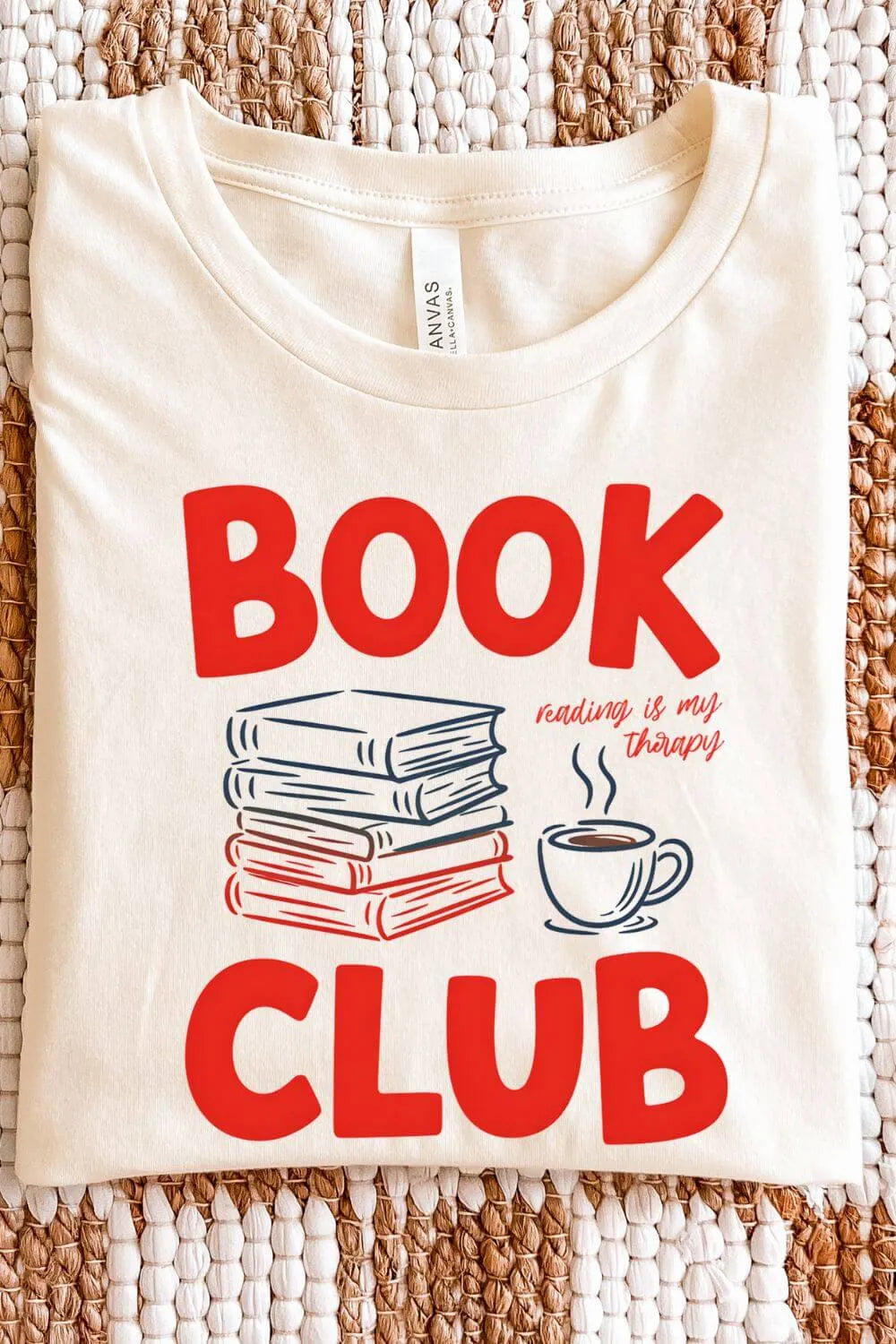 soft cotton natural beige  graphic t-shirt with bold red text reading “Book Club,” a stack of books, a coffee cup, and the phrase “Reading Is My Therapy” in a cozy bookish design, folded on textile surface