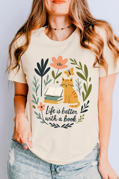 A soft cotton cream-colored t-shirt worn by a model, featuring a cute illustrated cat sitting beside stacked books with leafy florals and the phrase ‘Life Is Better With a Book.’