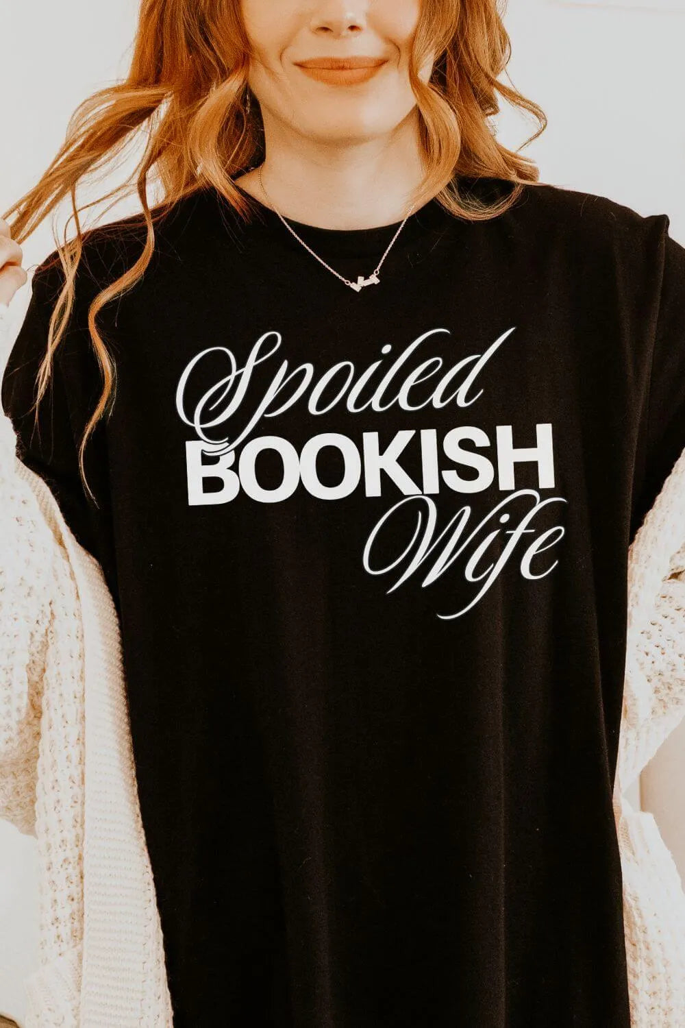 Woman wearing a black t-shirt with the phrase “Spoiled Bookish Wife” in elegant typography, a funny book lover shirt styled with a cozy cardigan and aesthetic outfit.