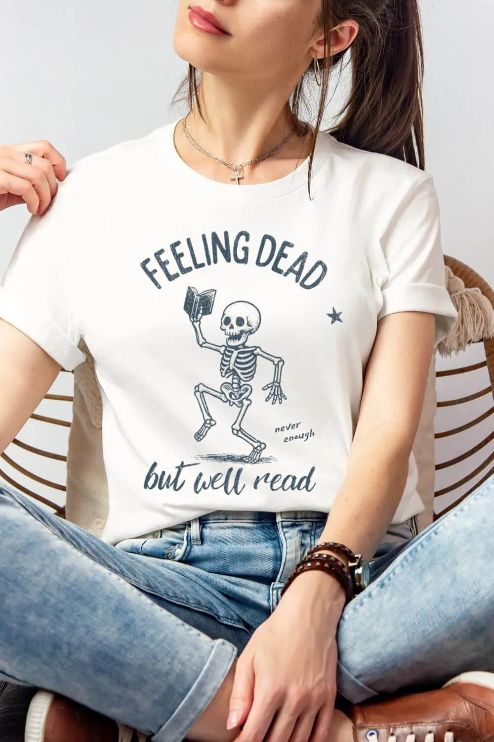 Person wearing a white soft cotton graphic t-shirt featuring a vintage-style skeleton holding a book with the text “Feeling Dead but Well Read,” styled casually with jeans for a dark academia reader outfit.