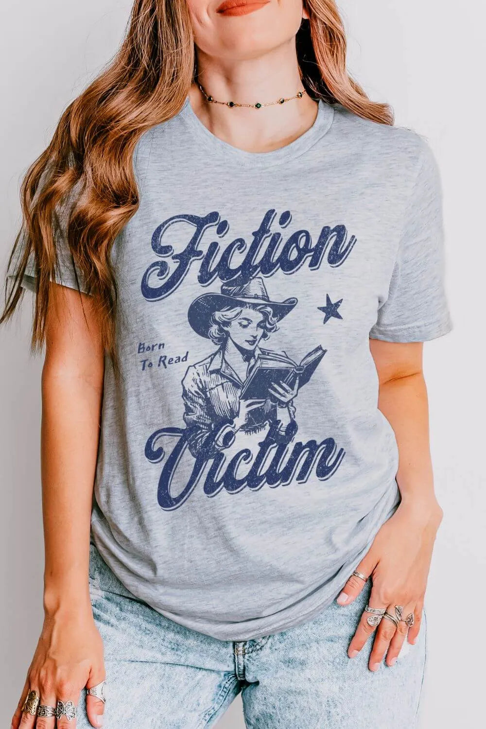 Athletic heather gray t-shirt worn by a model, featuring a vintage-style cowboy illustration of a  woman reading with the words ‘Fiction Victim’ and ‘Born to Read’ in distressed navy lettering