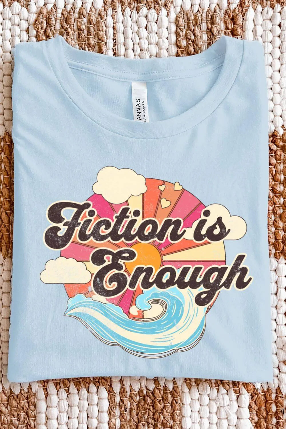 A folded light blue retro-style graphic t-shirt with colorful sunset and ocean wave artwork and bold vintage lettering that reads “Fiction Is Enough” for groovy aesthetic outfit