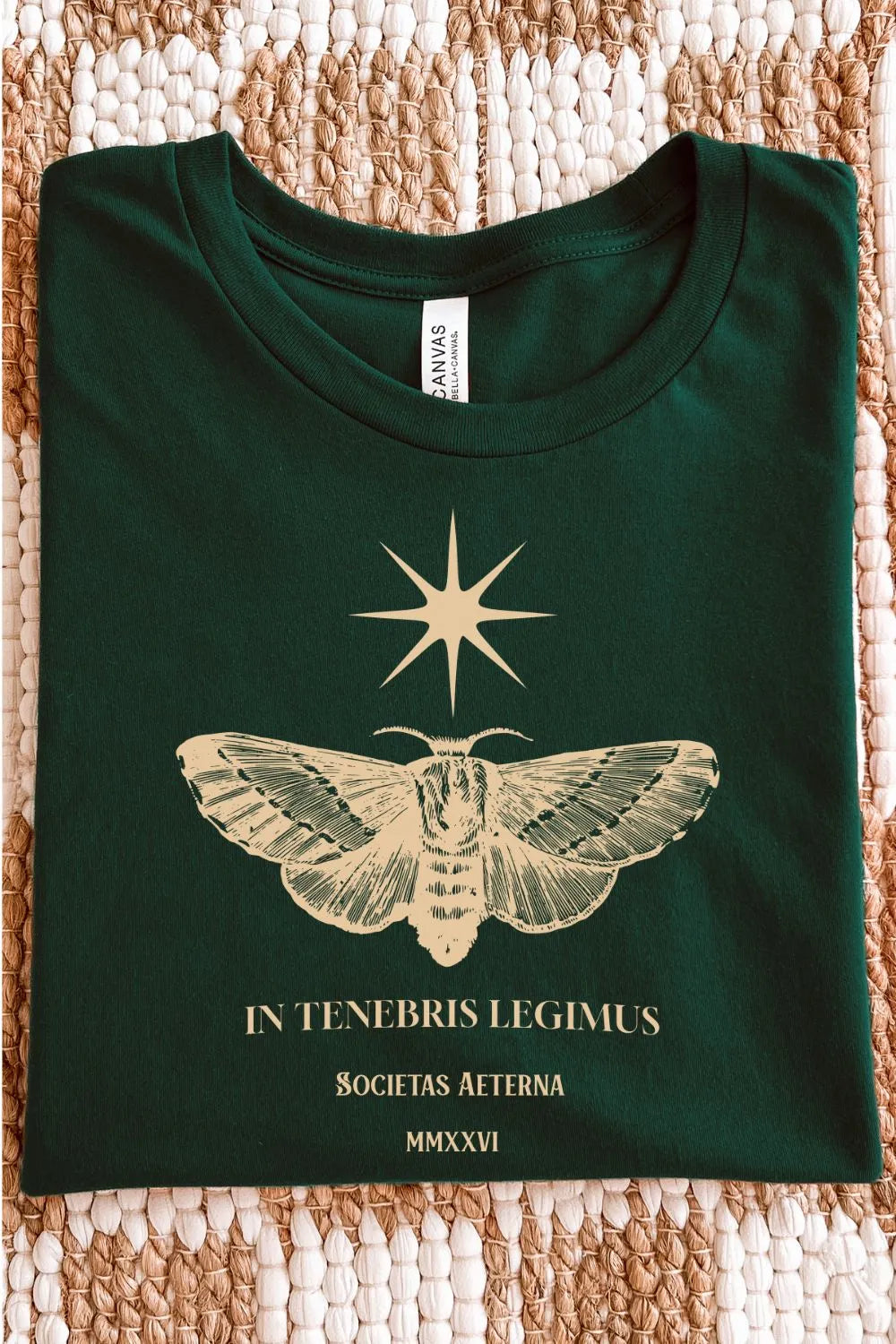A minimal literary tee for introverted book lovers and midnight readers featuring latin quote in golden typography and vintage moth illustration in forest green, folded on textile surface