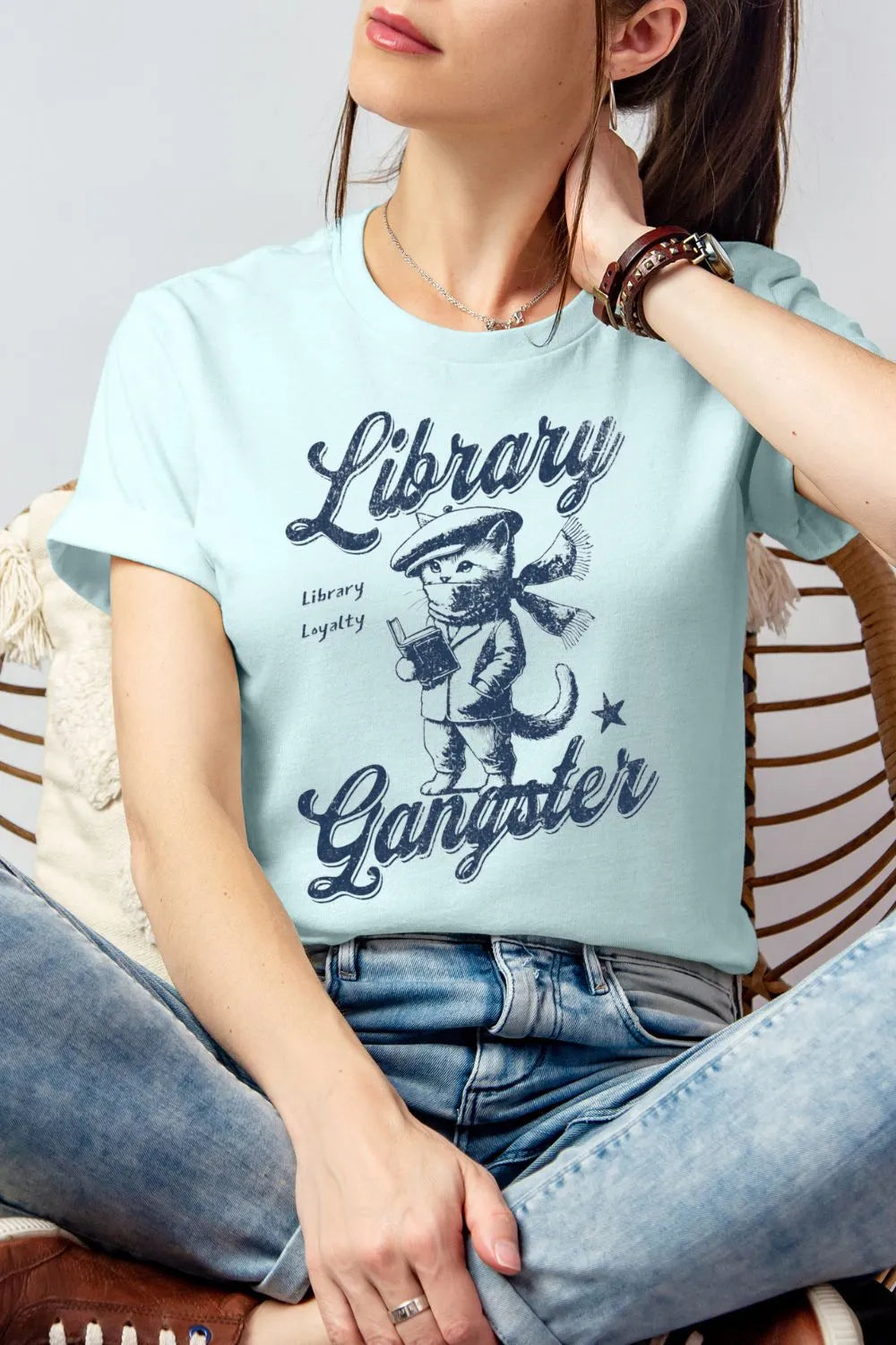 A woman wearing soft heather ice blue vintage-style t-shirt featuring a retro illustrated cat holding a book with the text “Library Gangster,” styled casually with jeans for a cozy reader outfit