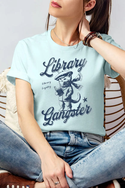 A woman wearing soft heather ice blue vintage-style t-shirt featuring a retro illustrated cat holding a book with the text “Library Gangster,” styled casually with jeans for a cozy reader outfit