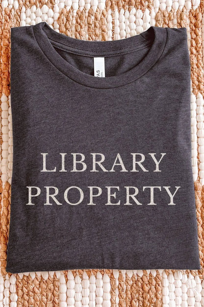 Folded dark gray heather  t-shirt with the phrase “Library Property” printed in vintage typography, a minimalist book lover shirt styled on a woven background for a cozy library aesthetic.
