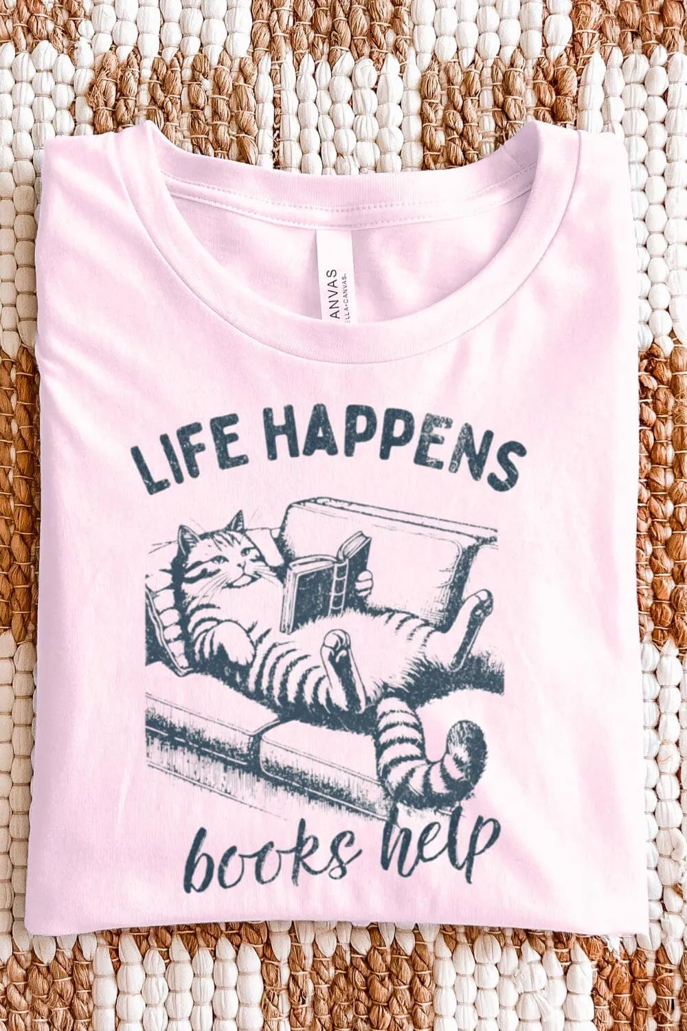 A soft pink t-shirt featuring a vintage-style illustration of a relaxed cat reading a book with the phrase “Life Happens, Books Help,” designed in a cozy bookish aesthetic