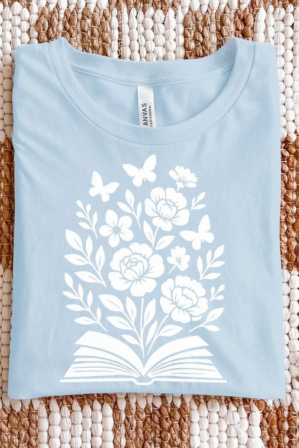 A light blue soft cotton  t-shirt with a white illustration of an open book blooming with flowers and butterflies, designed in a soft, nature-inspired bookish style.
