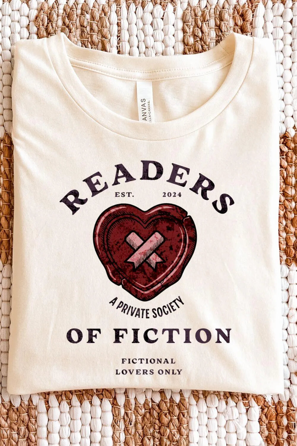 A  soft cotton cream vintage-style t-shirt with a distressed heart emblem and text reading “Readers of Fiction – A Private Society,” designed with classic typography and bookish humor.