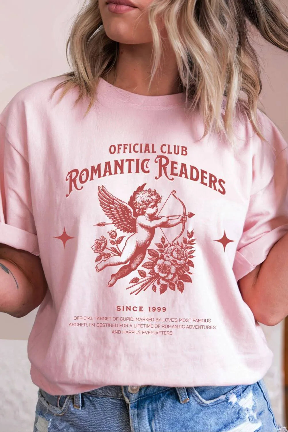 A soft pink t-shirt worn by a model, featuring a vintage red Cupid illustration with roses and the text ‘Official Club Romantic Readers,’ styled casually with denim